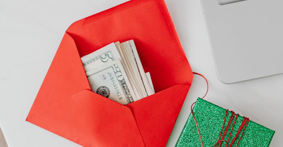 A red envelope containing cash beside a laptop and glittering green gift box on a white table.
